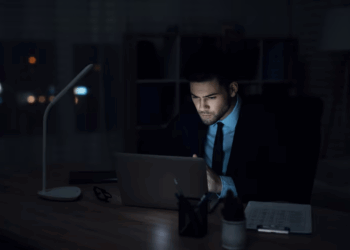 A man in a suit works on his laptop at night, illuminated by the screen's glow in a dimly lit room