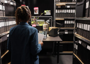 Police investigator holding crime matter file and analyzing evidence board in office late. African american woman law enforcement detective studying clues, working overtime at night