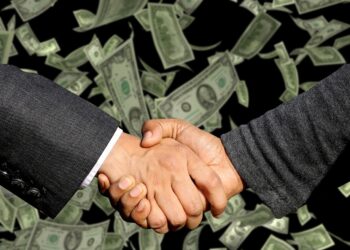 Two businessmen engage in a handshake above a substantial pile of money, representing a lucrative business deal