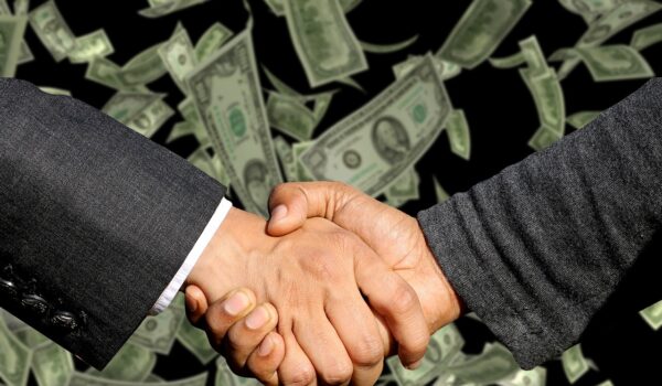 Two businessmen engage in a handshake above a substantial pile of money, representing a lucrative business deal