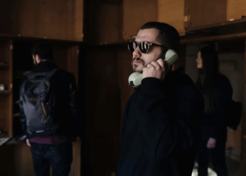 A man wearing sunglasses is talking on a phone while seated in a room