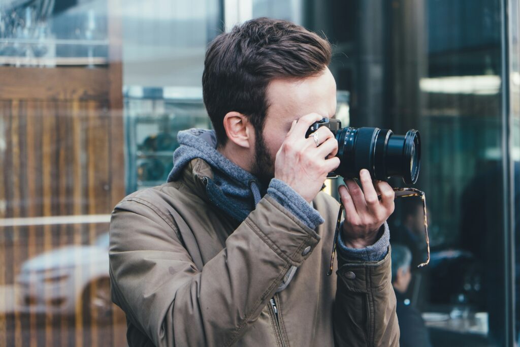 A person in a jacket and hoodie holds a camera up to their face, focusing intently as they take a photo outdoors