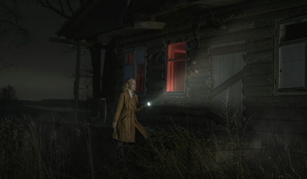 A woman with a flashlight approaches a dark, abandoned house with red-lit windows at night